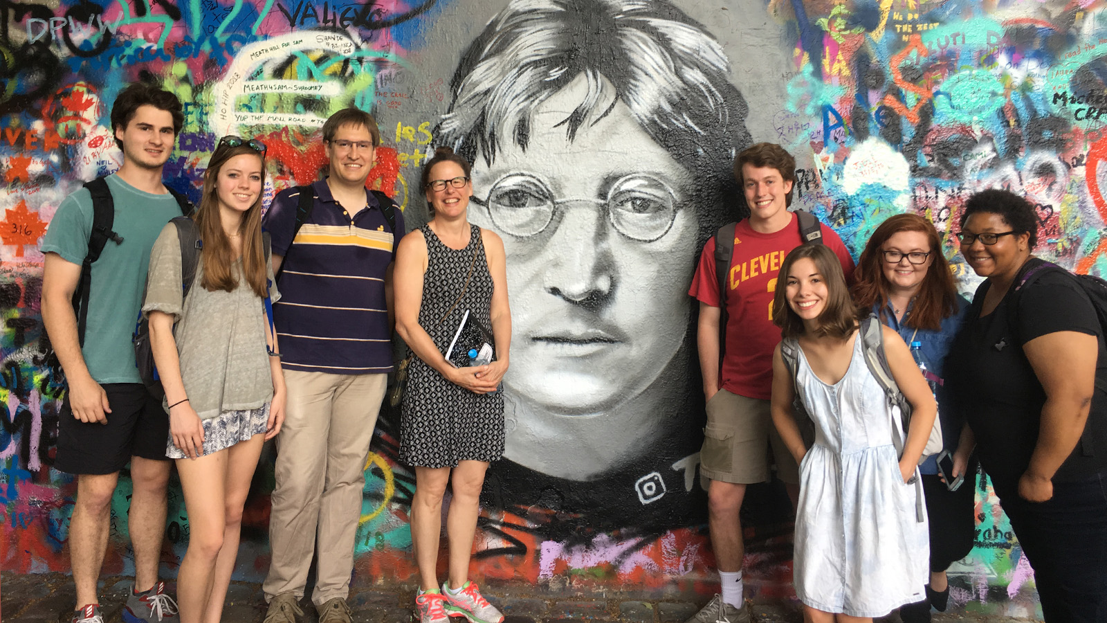 Students and faculty stand in front of the John Lennon wall.